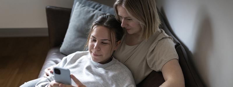 Två unga kvinnor sitter i soffa och tittar på mobil